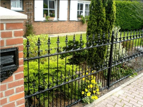 Svart dekorativt metallstaket framför en tegelfasad med grönskande trädgård, buskar, blommor och en brevlåda.